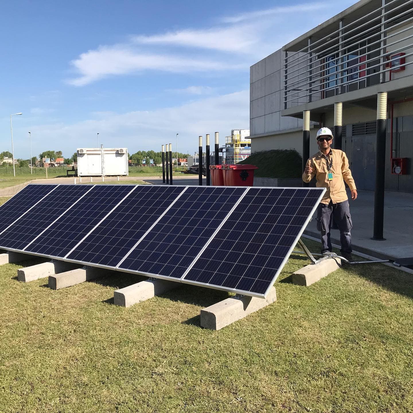 Matías con casco, con el pulgar arriba, junto a una fila de paneles solares apoyados sobre bases de hormigón en el pasto.